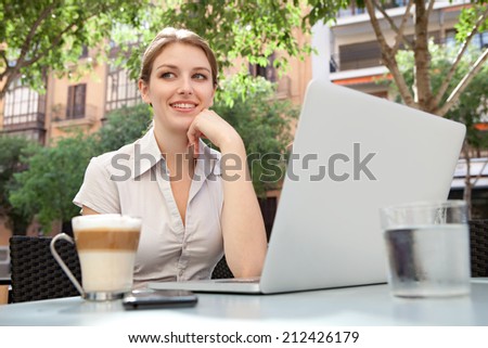 Close up portrait of a young professional business woman sitting at a cafe terrace shop drinking a coffee and using a laptop computer, thoughtful and smiling in a financial city district, outdoors.