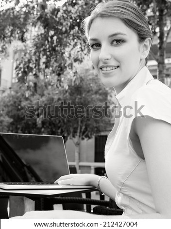 Black and white side view of an attractive young professional business woman sitting at a cafe terrace shop drinking coffee and using a laptop computer in a financial city, outdoors.