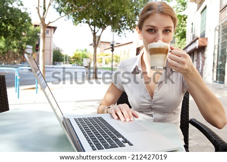Portrait of an attractive young professional business woman sitting at a cafe terrace shop drinking  a hot beverage coffee and using a laptop computer in a classic city, outdoors.