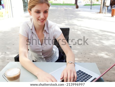 Beautiful professional young business woman sitting at a coffee shop drinking a beverage and using laptop computer in the financial city, smiling outdoors. Business people at work.