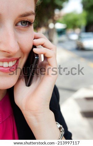 Close up half face portrait of a beautiful young business woman using a smartphone in the city to make a call and have a conversation, smiling in a classic street. Professional woman using technology.