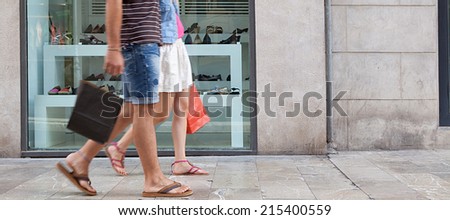Panoramic side view of a couple lower body section walking in shopping street with a shoe store window display, carrying shopping bags and spending money on holiday. Consumer lifestyle, outdoors.