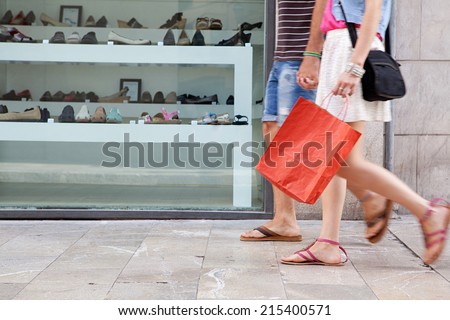 Side view of a young couple lower body section walking down a shopping street with a shoe store window display, carrying shopping bags and spending money on holiday. Consumer lifestyle, outdoors.