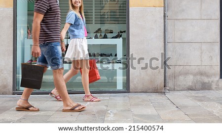 Side view of a young couple holding hands walking in shopping street with a shoe store window display, carrying shopping bags and spending money on holiday. Consumer lifestyle, outdoors.