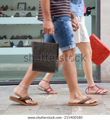 Side view of a young couple lower body section walking down a shopping street with a shoe store window display, carrying shopping bags and spending money on holiday. Consumer lifestyle, outdoors.