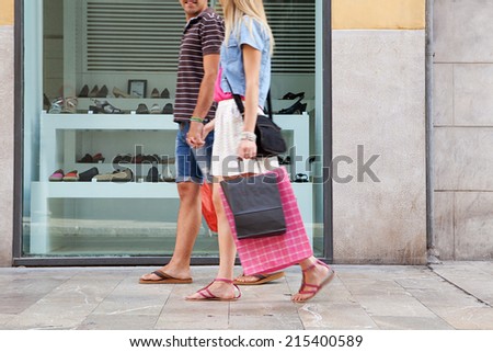 Side view of a young couple lower body section walking in shopping street with a shoe store window display, carrying shopping bags and spending money on holiday. Consumer lifestyle, outdoors.