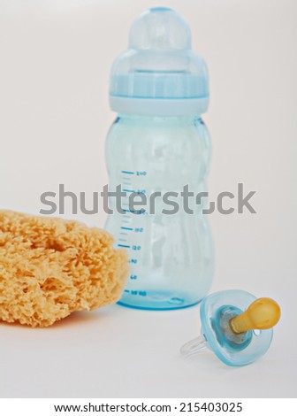 Close up still life of a baby dummy and feeding bottle together with a bath washing sponge on a white background. Detail view of a nursery new born items.