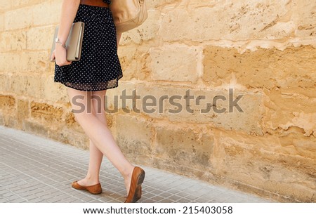 Rear view of a professional young businesswoman legs walking along an old stone building wall while commuting to work and carrying a folder with paperwork, outdoors.