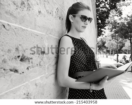 Black and white portrait of a thoughtful professional business woman holding and reading diary while leaning on an old stone building wall in a classic city, outdoors. Smart professional young woman.