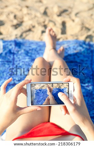 Close up of a young and attractive woman tourist hands together holding a modern technology smartphone and taking a selfie picture of her own legs while laying down on a sandy beach on holiday.