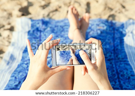 Close up of a young and attractive woman tourist hands together holding a modern technology smartphone and taking a selfie picture of her own legs while laying down on a sandy beach on holiday.