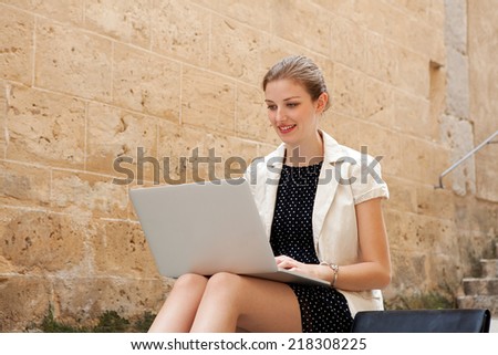 Young professional business woman sitting on the steps of an old stone building using a laptop computer working outdoors, smiling. Connectivity and wireless internet browsing.