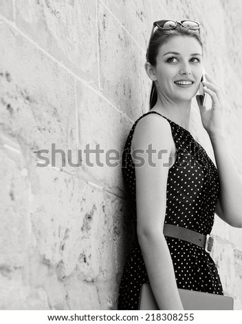 Black and white portrait of a beautiful young professional businesswoman using a smart phone technology to make a phone call, leaning on a textured stone wall in a financial city. Lifestyle outdoors.