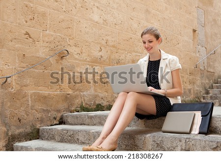Young professional business woman sitting on the steps of an old stone building using a laptop computer working outdoors, smiling. Connectivity and wireless internet browsing.