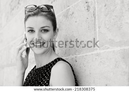 Black and white portrait of beautiful young professional businesswoman using a smart phone technology to make a phone call, leaning on a textured stone wall in a financial city. Lifestyle outdoors.