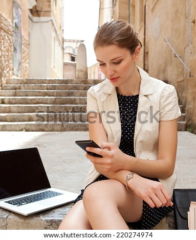 Attractive young professional business woman sitting on the steps of an old stone building using a smartphone and a laptop computer working outdoors. Connectivity and wireless internet browsing.