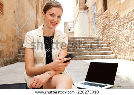 Attractive young business woman using smartphone and laptop computer while sitting on a stone steps street in a classic city and smiling outdoors. Professional corporate people and technology.