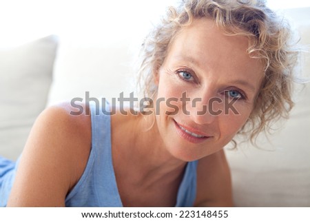Beauty portrait of an attractive middle aged healthy woman relaxing on a white sofa at home, looking at the camera smiling. Interior home living and well being lifestyle.