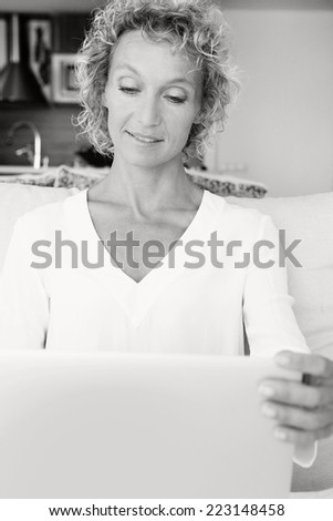 Black and white portrait of a beautiful professional mature woman sitting on a white sofa at home by the kitchen, using a laptop computer and working, smiling. Home living technology lifestyle.