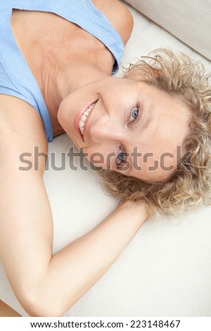 Over head close up beauty portrait of an attractive middle aged healthy woman relaxing on a white sofa at home, looking at the camera smiling. Interior home living and lifestyle.