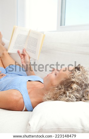 Over head close up beauty portrait of an attractive middle aged healthy woman relaxing on a white sofa at home, reading a book having a break. Interior home living and well being lifestyle.