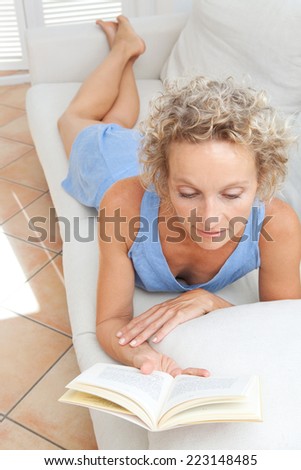 Beauty portrait of an attractive middle aged healthy woman relaxing on a white sofa at home, laying down and reading a book having a break. Interior home living and well being lifestyle.
