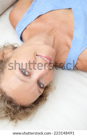 Over head close up beauty portrait of an attractive middle aged healthy woman relaxing on a white sofa at home, looking at the camera smiling. Interior home living and well being lifestyle.