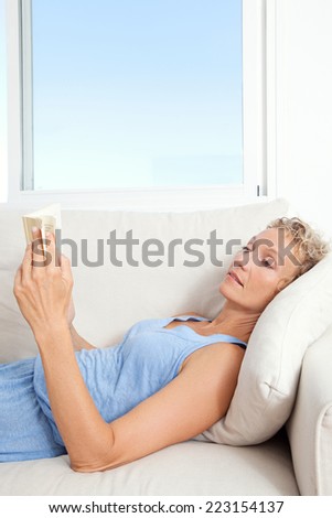 Side portrait of a beautiful middle age healthy woman laying down on a white sofa at home, holding and reading a book indoors. Woman relaxing and home living lifestyle.