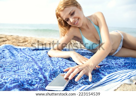 Portrait of an attractive teenager girl laying down relaxing on holiday, using a smartphone device on vacation laying down on a beach towel on the sand. People travel technology.