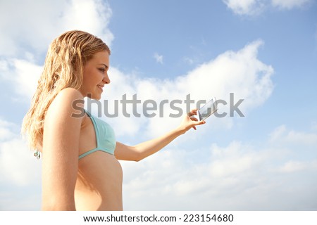 Portrait of an attractive teenager girl standing taking pictures on her mobile phone on holiday, holding a smartphone device taking photos, on vacation on sunny blue sky. People travel technology.