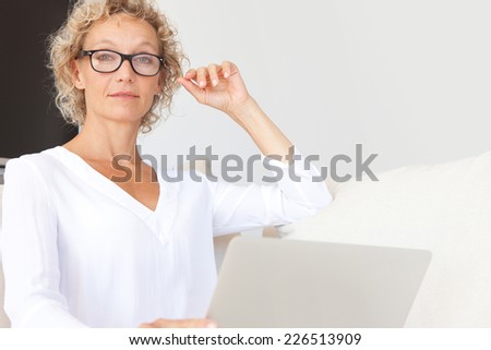 Portrait of a confident mature professional woman sitting on a white sofa at home using a laptop computer to work, indoors. Aspirational businesswoman wearing glasses smiling and working from home.