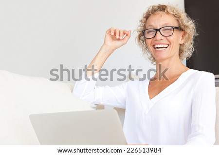 Portrait of a confident mature professional woman sitting on a white sofa at home using a laptop computer to work, joyfully laughing indoors. Businesswoman wearing glasses and working from home.