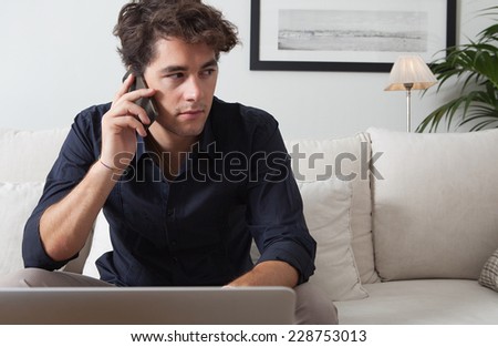 Close up portrait of an attractive professional businessman sitting on a coach in his home living room, working on his computer and making a phone call with a smartphone, indoors. Working home office.