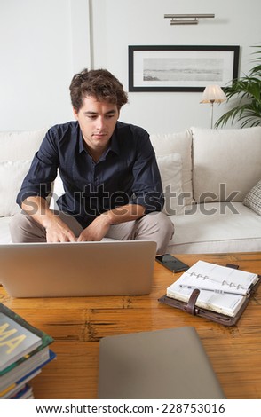 Attractive young professional business man sitting on a white coach in his home living room, working on his laptop computer and looking at folders and paperwork, indoors. Working from home office.