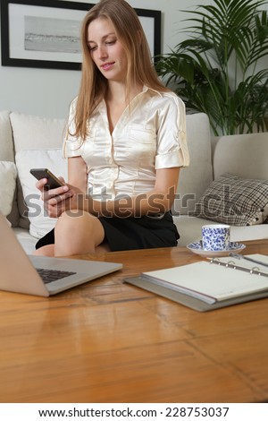 Portrait of an attractive young professional business woman sitting on a white sofa at home, using a smartphone and laptop computer to work, home interior. Working from home office with technology.
