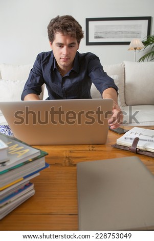 Attractive young professional business man sitting on a white coach in his home living room, working on his laptop computer and looking at folders and paperwork, indoors. Working from home office.