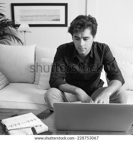 Black and white portrait of a young professional businessman sitting on a white coach at home, working on his laptop computer and looking at folders and paperwork, indoors. Working from home office.