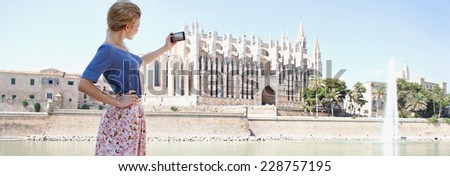 Panoramic side view of a tourist girl standing and using a smartphone to take photos of a cathedral in a destination city on vacation. Holiday maker taking pictures with smart phone, screen showing.