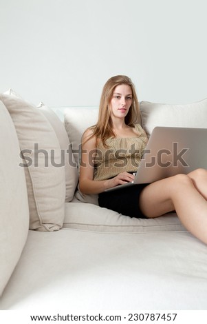 Attractive professional business woman using laptop computer to browse the internet on line, sitting on a spacious white wall living room with a comfortable white sofa, home interior. Space for type.