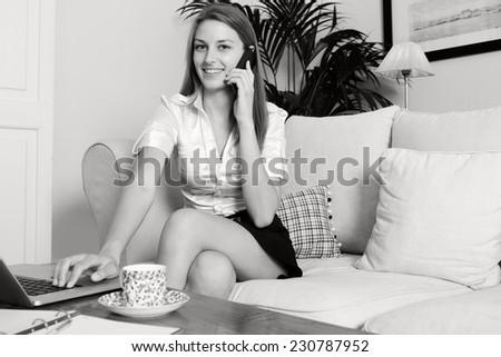 Black and white portrait of an young professional business woman sitting on a white sofa at home making a call on a smartphone, working on laptop computer, drinking a coffee smiling, home interior.