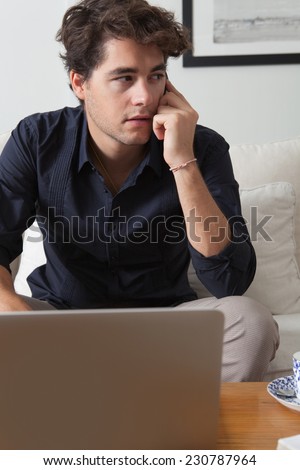 Attractive professional businessman sitting on a coach in his home living room, working on his laptop computer and making a phone call with a smartphone, indoors. Working from home office.