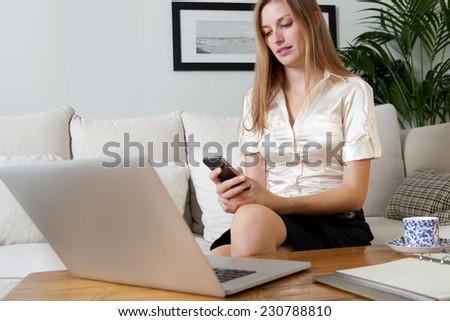Portrait of an attractive young professional business woman sitting on a white sofa at home, using a smartphone and laptop computer to work, home interior. Working from home office with technology.