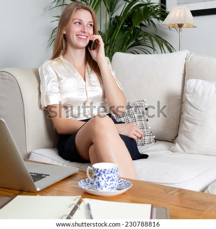 Portrait of beautiful young professional business woman sitting on a white sofa at home, making a call with smartphone and laptop computer to work, home interior. Working from home with technology.