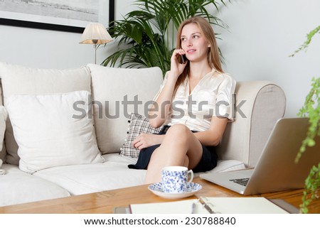 Portrait of an attractive young professional business woman sitting on a white sofa at home making a call on a smartphone, working using a laptop computer and drinking a coffee, home interior.