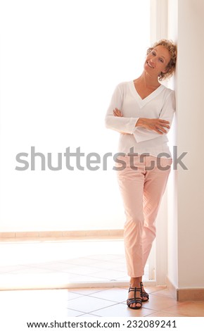 Smart professional business woman relaxing leaning on a wall near a glass door with sunny light, relaxing and enjoying her aspirational lifestyle, indoors. Home interior with businesswoman standing.