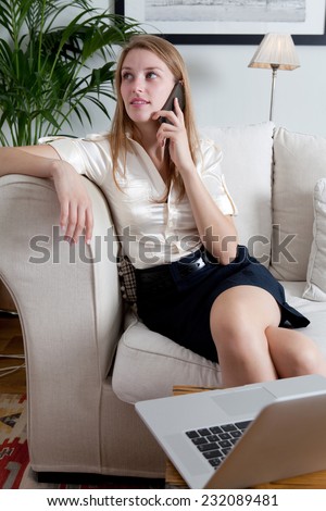Attractive professional businesswoman sitting on a coach at home living room, working on a laptop computer and making a phone call with a smartphone, indoors. Working from home office.