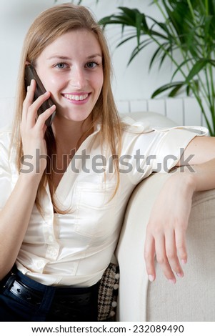 Attractive professional businesswoman sitting on a coach at home living room, making a phone call with a smartphone, joyfully smiling indoors. Working from home office.