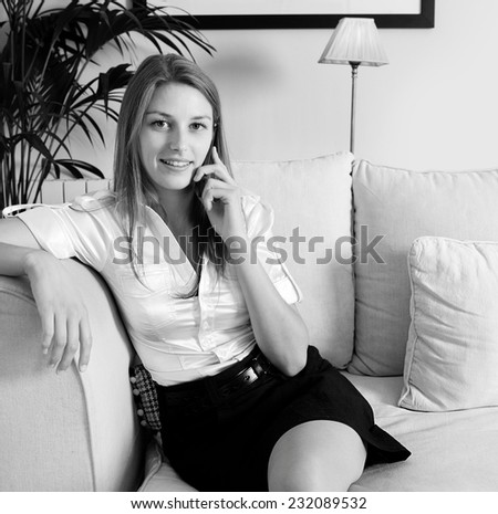 Black and white portrait of attractive young professional business woman sitting on a white coach at home living room smiling, having a telephone conversation on her spartphone device, indoors.