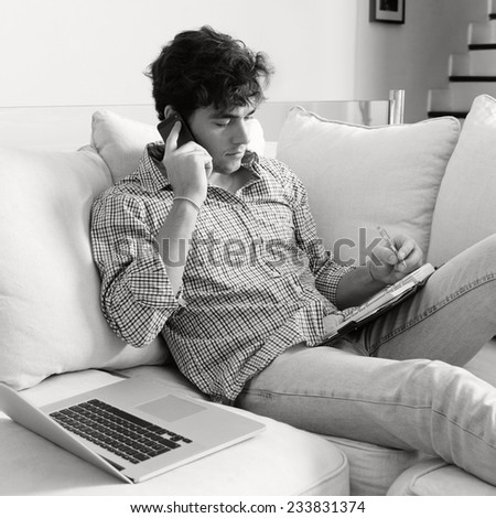 Black and white portrait of a young business man sitting on a white sofa at home, making a call on his smartphone and using a laptop computer. Professional man working from home, sitting on sofa.