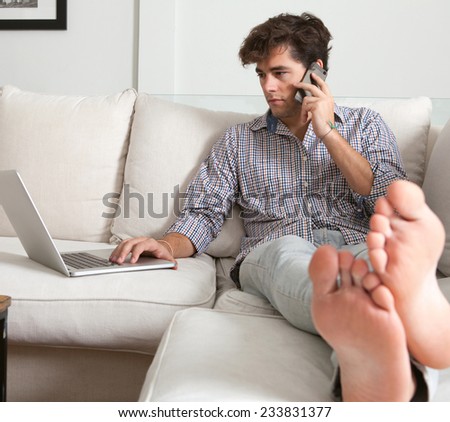 Young attractive business man sitting on a white sofa in his home living room, making a call on his smartphone and using a laptop computer. Professional businessman working from home, sitting on sofa.
